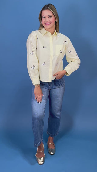 A woman modeling a pale yellow long-sleeve button-up shirt with small Mallard duck prints, paired with blue jeans and metallic shoes, standing against a solid blue studio backdrop.