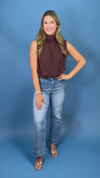 A woman with blonde-highlighted hair wearing a sleeveless high-neck brown top and blue jeans
