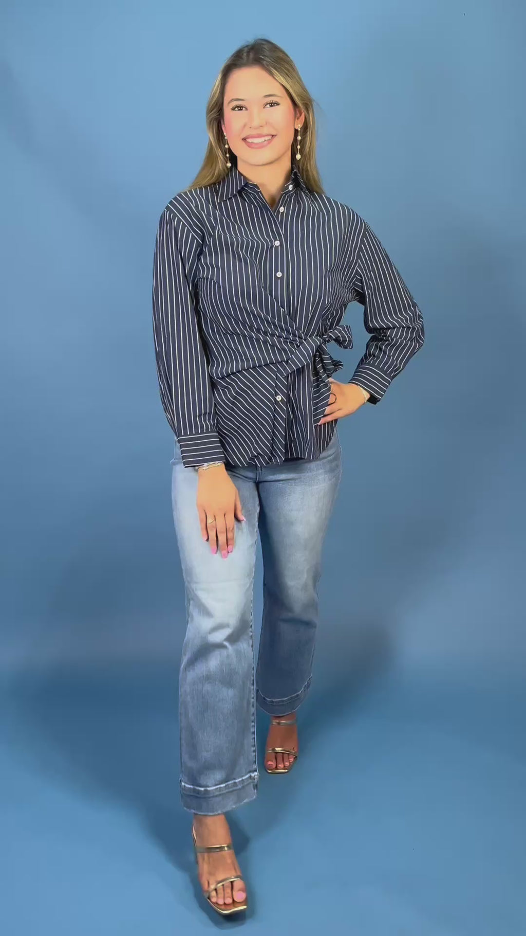 A woman modeling a navy blue and white vertically striped long-sleeve button-up shirt with a tie waist, paired with light-wash blue jean