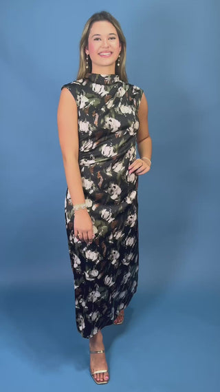 A woman with long blonde-highlighted hair wearing a sleeveless black dress with a white and green floral print