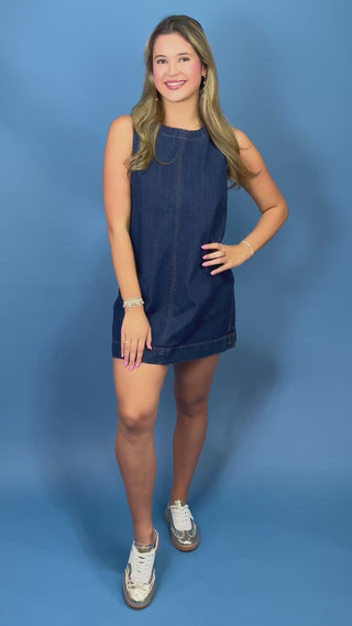 A smiling young woman with blonde hair models a sleeveless dark blue denim mini dress 