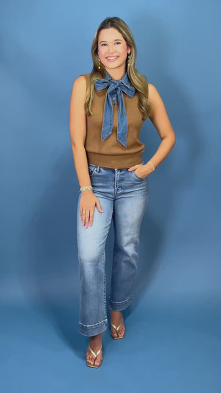 A woman modeling a sleeveless brown knit sweater vest with a large blue denim bow at the neckline, paired with light-wash blue jeans