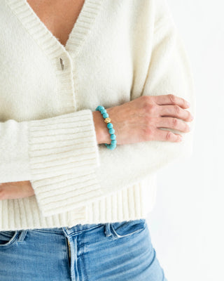 Person wearing a white sweater and blue jeans with a turquoise bracelet on a white background