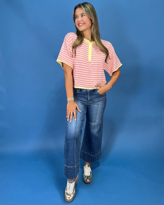 A woman modeling a pink and yellow striped short-sleeve henley top with yellow trim, paired with wide-leg blue jeans and whit