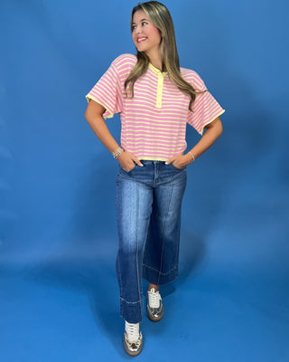 A woman modeling a pink and yellow striped short-sleeve henley top with yellow trim, paired with wide-leg blue jeans and whi