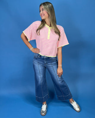 A woman modeling a pink and yellow striped short-sleeve henley top with yellow trim, paired with wide-leg blue jeans and whit