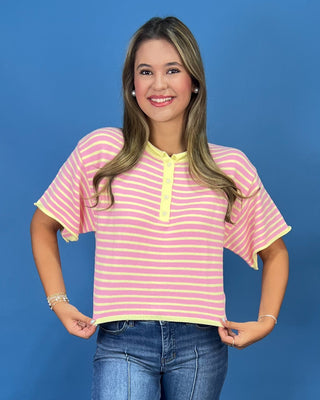 A woman modeling a pink and yellow striped short-sleeve henley top with yellow trim, paired with wide-leg blue jeans and whit
