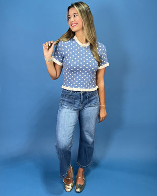 a light blue short-sleeve top with white polka dots and white ruffled trim, paired with blue jeans, standing against a soli