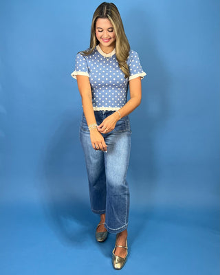 A woman modeling a blue and white polka dot short-sleeve crop top with cream ruffle trim, paired with blue jeans and metalli