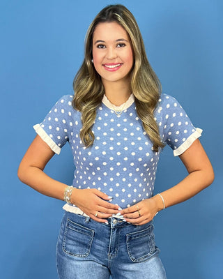 a light blue short-sleeve top with white polka dots and white ruffled trim, paired with blue jeans, standing against a soli