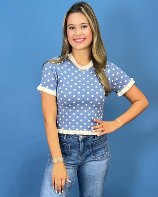 a light blue short-sleeve top with white polka dots and white ruffled trim, paired with blue jeans, standing against a soli