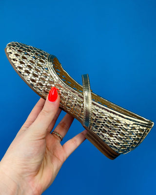 A hand with red nail polish holds a metallic gold flat shoe with a textured, woven-like pattern and a single strap across the