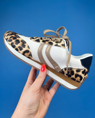 A hand with red nail polish holds a low-top sneaker featuring white panels, beige side stripes, and leopard print over