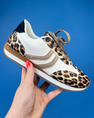 A hand with red nail polish holds a low-top sneaker featuring a leopard print pattern on the toe, heel, and laces, combined w