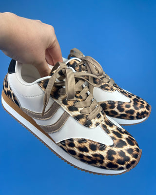 A pair of low-top sneakers with a leopard print and white color-blocked design, held by a hand against a solid blue backgrou