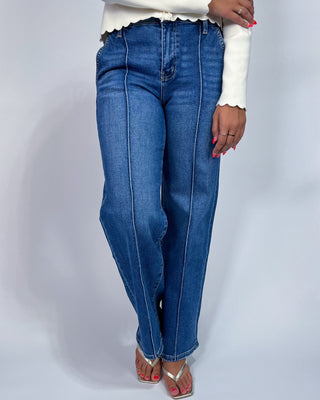 A person wearing medium-wash blue jeans with vertical contrast stitching, styled with a white long-sleeve top and metallic sa