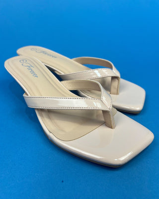 A pair of glossy beige patent leather thong sandals with square toes and low block heels, displayed on a vibrant blue backgr