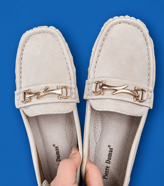 A pair of light beige suede penny loafers with gold-tone horsebit hardware, held open by a hand against a solid blue backgrou