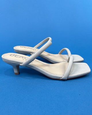 A pair of light gray suede-like heeled sandals with square toes and thin wrap-around straps, displayed on a solid blue b