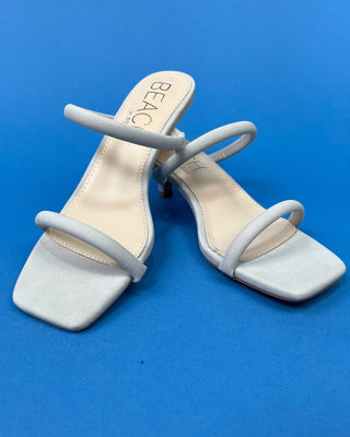 A pair of light gray suede-like heeled sandals with square toes and thin wrap-around straps, displayed on a solid blue b