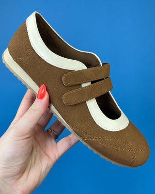 A brown suede slip-on shoe with cream-colored trim and double hook-and-loop straps, held by a hand with red nail polish agai