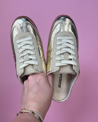 Metallic gold lace-up sneakers with a shiny textured finish, cream accents, white laces, and gum rubber soles.