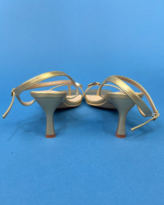 A pair of women's high-heeled sandals shown from the back, featuring metallic gold straps and a light beige footbed, s