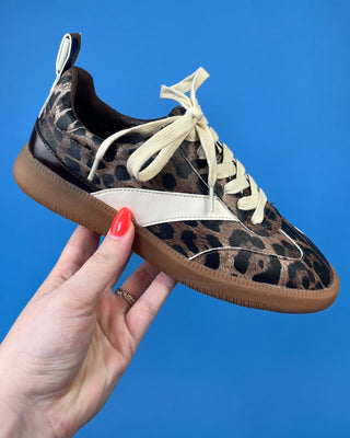A leopard print sneaker with cream laces and a white side stripe, held by a hand with red nail polish against a solid