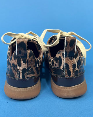 A pair of low-top sneakers shown from the back, featuring a brown and black leopard print upper, cream-colored laces, and t