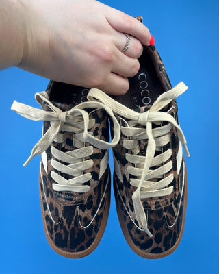 A pair of low-top sneakers with a brown and black leopard print pattern, cream-colored laces, and white side stripes, held by