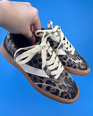 A pair of low-top sneakers with a brown and black leopard print pattern, cream-colored laces, and a white side stripe, hel.