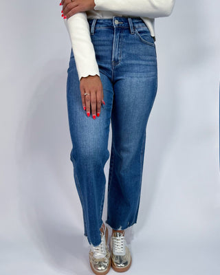 A person wearing medium-wash blue jeans with a white long-sleeve top and metallic silver sneakers, shown from the waist down