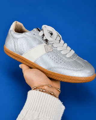 A metallic silver low-top sneaker with a white side stripe, white laces, and a brown gum sole, held up by a hand against a s