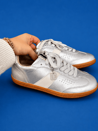 A pair of metallic silver low-top sneakers with white laces and gum soles, held by a hand wearing a white ribbed sweat