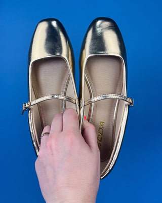 Metallic gold Mary Jane flats with a silver buckle strap, held by a hand with red nail polish against a solid blue background