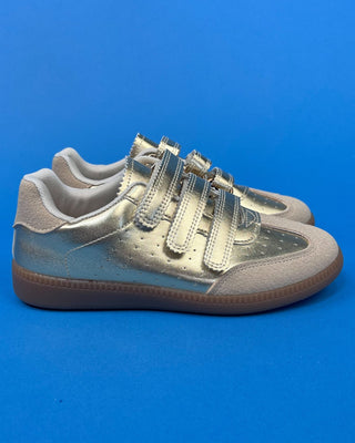 A pair of metallic silver low-top sneakers with three hook-and-loop straps and brown soles, set against a solid blue backgro