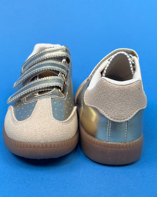 A pair of low-top sneakers shown from the back, featuring beige suede and metallic silver panels with a gold accent o