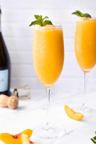 Two glasses of orange cocktail with peach slices and mint leaves, accompanied by a bottle of champagne on a light background.