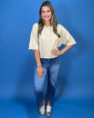 A woman modeling a cream-colored short-sleeve button-up shirt with blue jeans and silver sneakers,