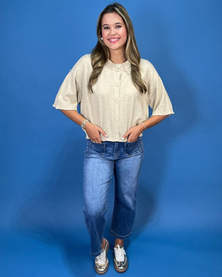 A woman modeling a short-sleeve cream linen button-up shirt with blue jeans and white sneakers, standing against