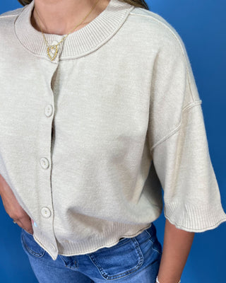 A close-up of a person wearing a light cream knit cardigan with a round collar, three-quarter sleeves, and front buttons, pa