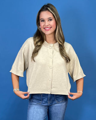 A woman modeling a short-sleeve cream knit cardigan with a button-down front, paired with blue jeans, against a solid blu