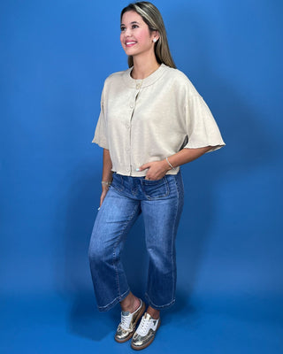A woman modeling a short-sleeve cream-colored button-up top with blue cropped jeans and metallic sneakers,