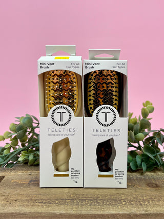 Two Teleties mini vent brush packages on a wooden surface with a pink background and greenery.