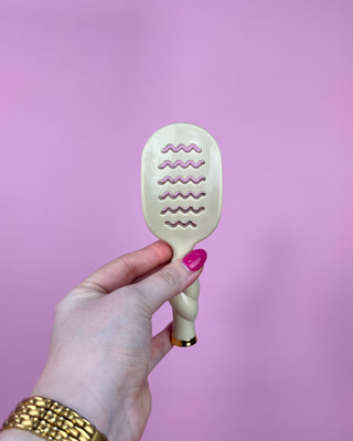 Hand holding a paddle brush with wavy pattern against a pink background