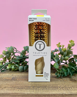 Teleties mini vent brush packaging on a wooden surface with a pink background and green leaves.