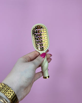 Hand holding a hairbrush with a gold and white design against a pink background