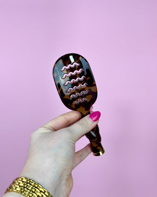 Tortoiseshell hairbrush held by a hand with pink nail polish against a pink background