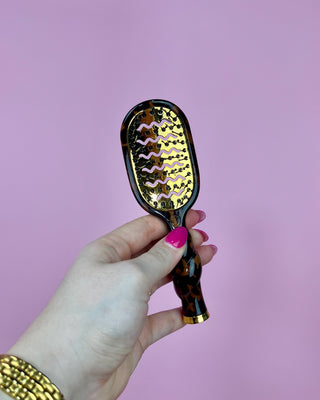 Hand holding a hairbrush with a leopard print handle against a pink background
