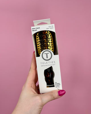Hand holding a Teleties mini vent brush packaging against a pink background